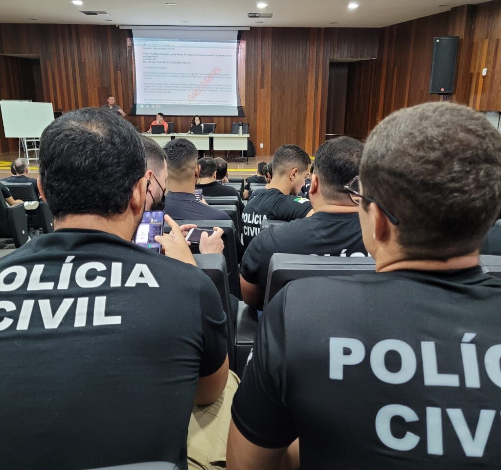 Imagem mostras homensa com camisetas pretas com o nome "Polícia Civil" em branco. Estão sentado em cadeiras de um auditório.