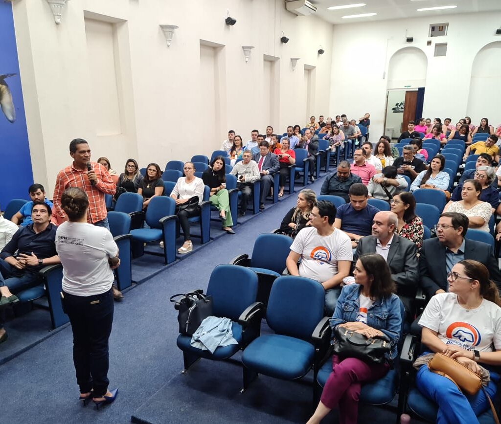 Imagem de um auditório com várias pessoas sentadas e uma mulher ao centro, de frente para a plateia.