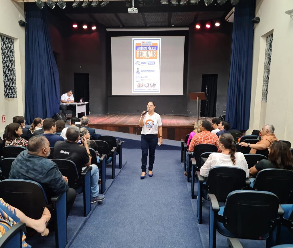 Imagem de um auditório repleto de pessoas e uma mulher, ao centro, falando ao microfone.