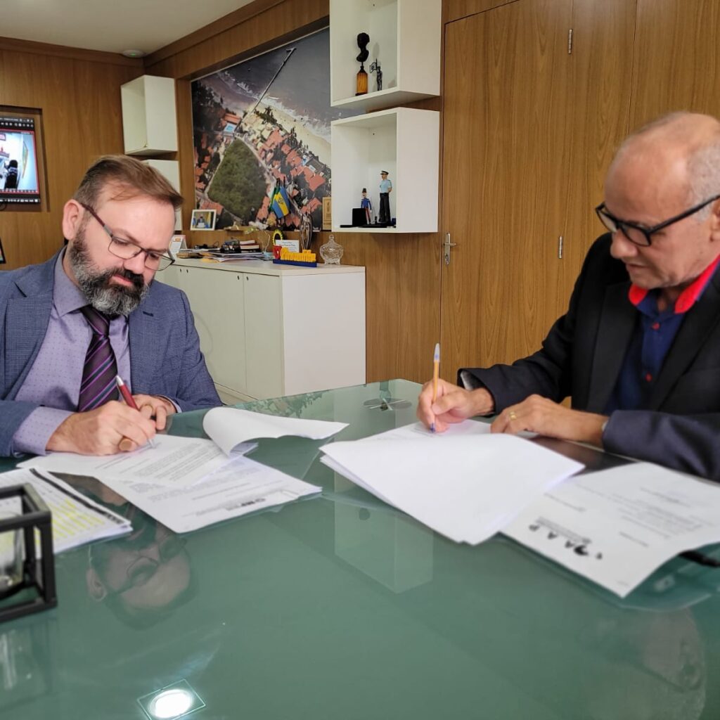 Imagem de dois homens sentados à mesa, cada um assinando um documento.