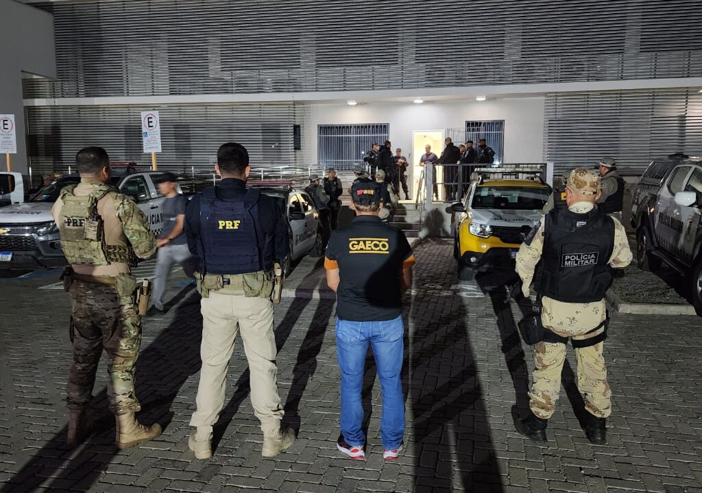 Imagem de agentes do Gaeco e policiais rodoviários federais e militares, de costas, em primeiro plano. Ao fundo, viaturas e mais policiais em frente a um prédio.