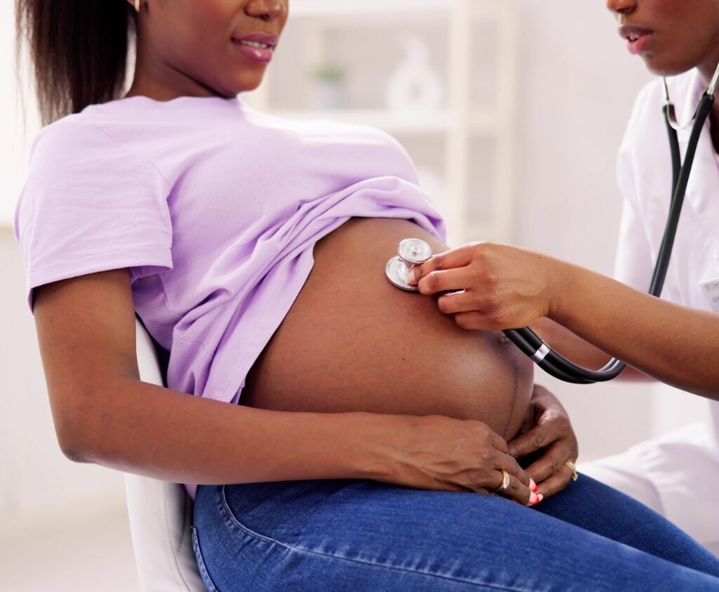 Imagem de uma mulher negra, grávida, sentada em uma cadeira e uma profissional de saúde fazendo a ausculta do bebê.