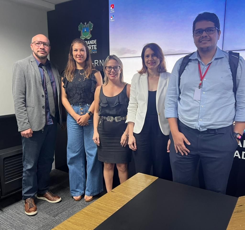 Imagem de cinco pessoas, sendo dois homens e três mulheres, todos em pé, em uma sala, posando para a foto.