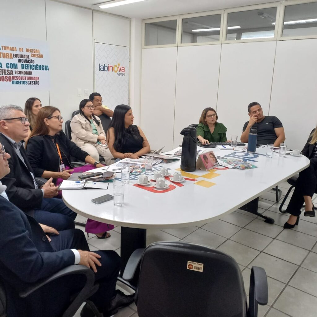 Imagem com várias pessoas sentadas ao redor de uma mesa oval, em uma sala, participando de uma reunião.