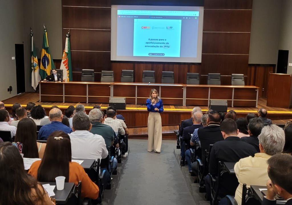 Imagem de uma mulher falando para uma plateia, em um auditório com várias pessoas.