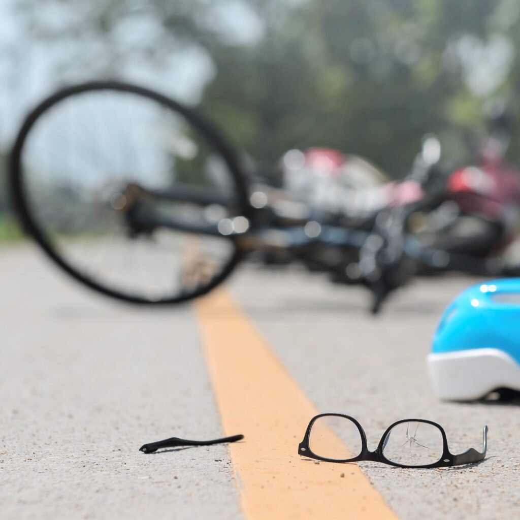 Imagem de bicicleta deitada em uma via de trânsito com capacete, óculos e itens pessoais espalhados após o acidente.