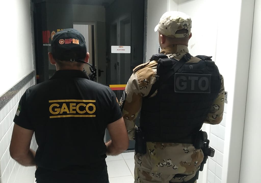 Imagem de um agente do Gaeco e de um policial militar, ambos de costas e em pé, em frente a uma porta de vidro.