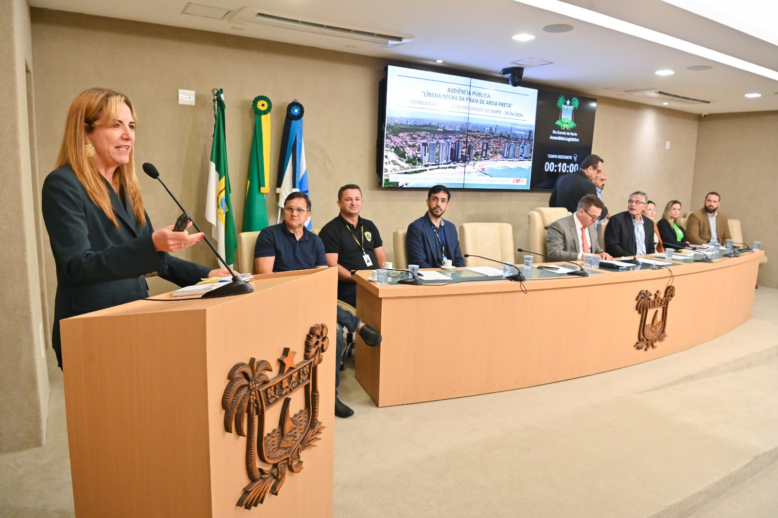 Foto mostrando do lado esquerdo a promotora falando em uma tribuna e ao lado direito a mesa de honra da solenidade.