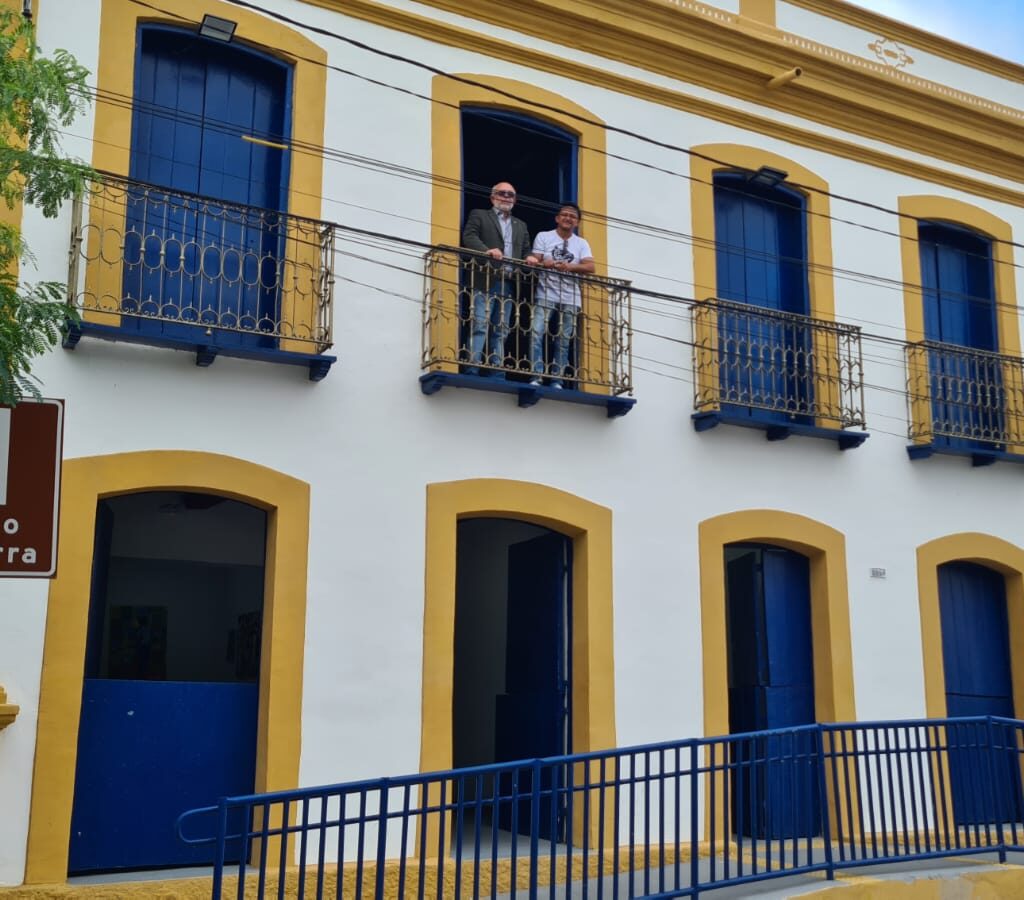 Imagem que mostas um sobrado de dois andares pintado de branco, com portas azuis e molduras das portas amarelas, tem dois homnes em uma das sacadas no andar superior.