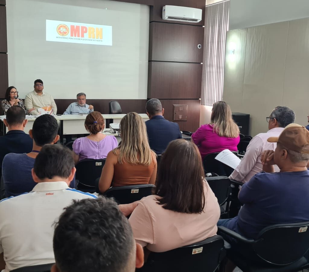 Foto de um auditório, com pessoas de costas,sentadas, em primeiro plano e ao fundo os promotores de Justiça sentados em uma meda, de frente para a plateia