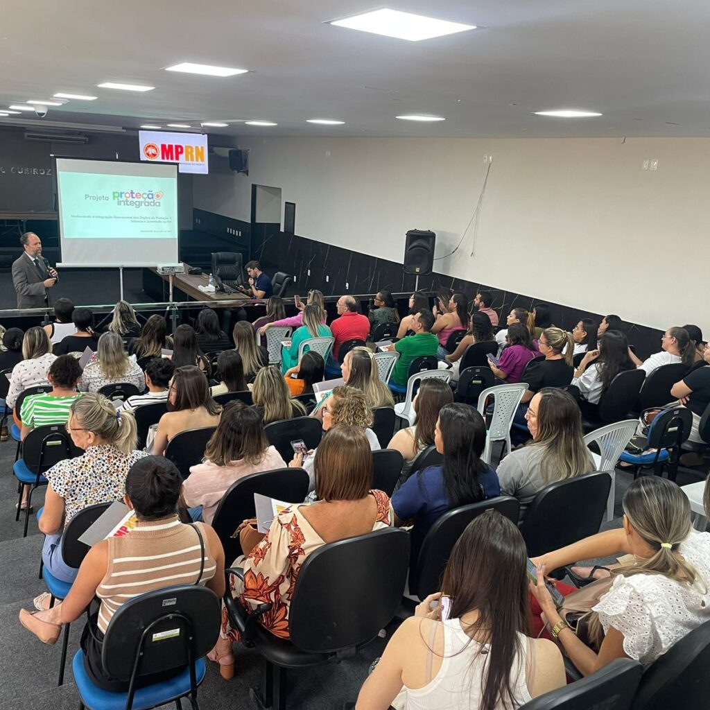 Imagem mostra um auditório da perspectiva de quem está atrás de todas as fileiras de cadeiras, todas ocupadas por pessoas. Ao fundo, perto do palco tem um banner com o slogan do MPRN e um homem branco em pé falando para o público.