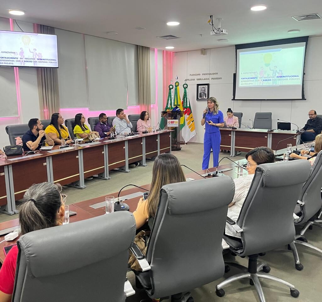 Imagem mostra um plenario com pessoas sentadas às mesas e uma mulher branca, vestida de azul, com um microfone, falando para os presente