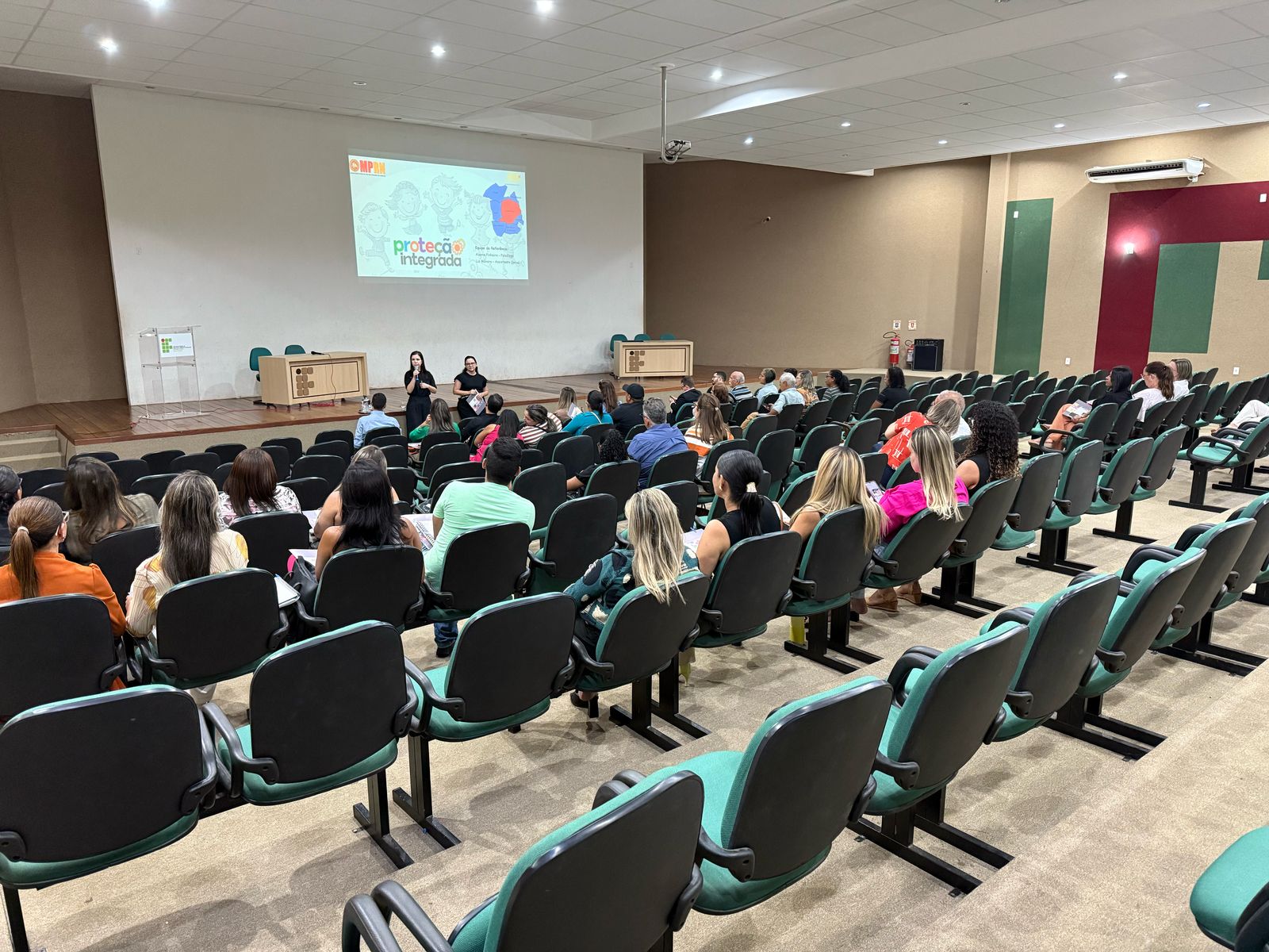 Imagem mostra auditorio com pessoas e apresentacao do projeto protecao integrada no datashow